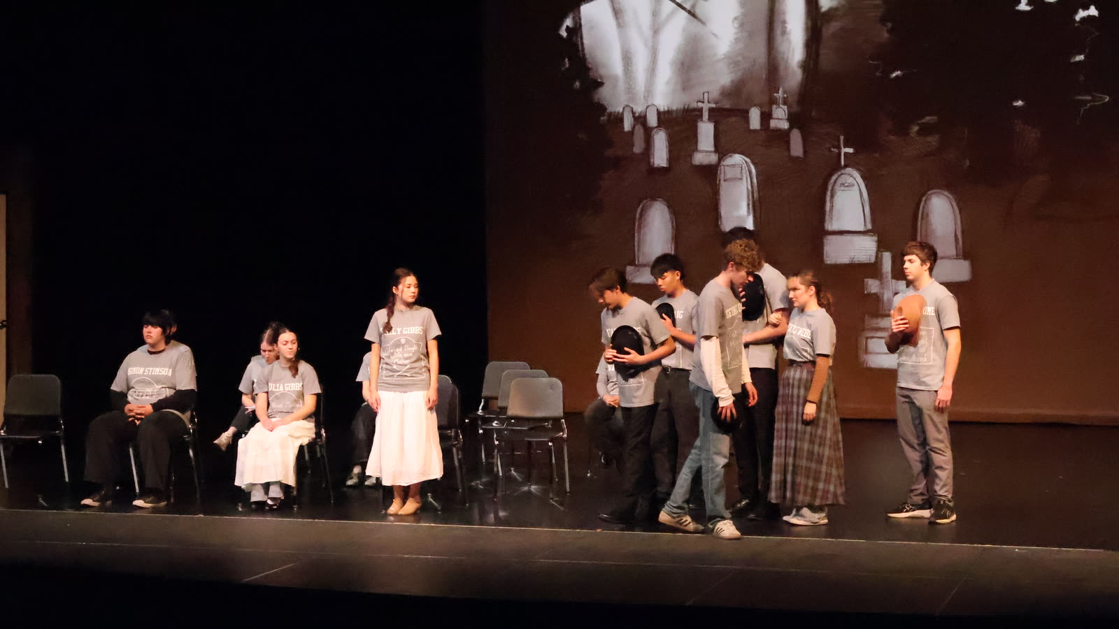 Drama students in a quiet ensemble scene before a painted graveyard backdrop